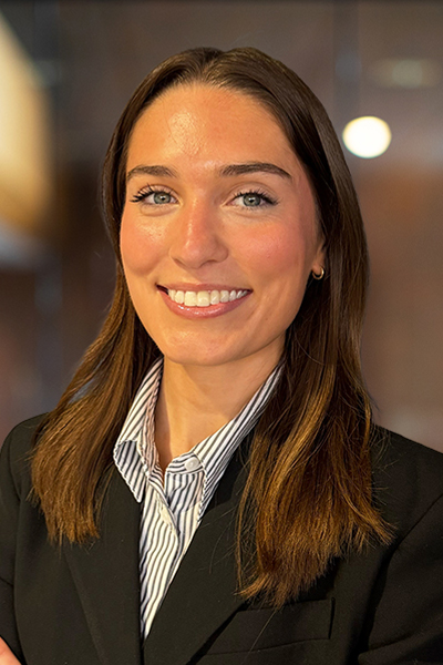 Portrait of a smiling woman with brown hair wearing a black blazer and striped collared shirt, in a professional indoor setting.